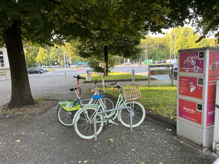 zwei Fahrräder an einem Fahrradständer unter einem Baum, rechts eine Ladesäule von Enercity