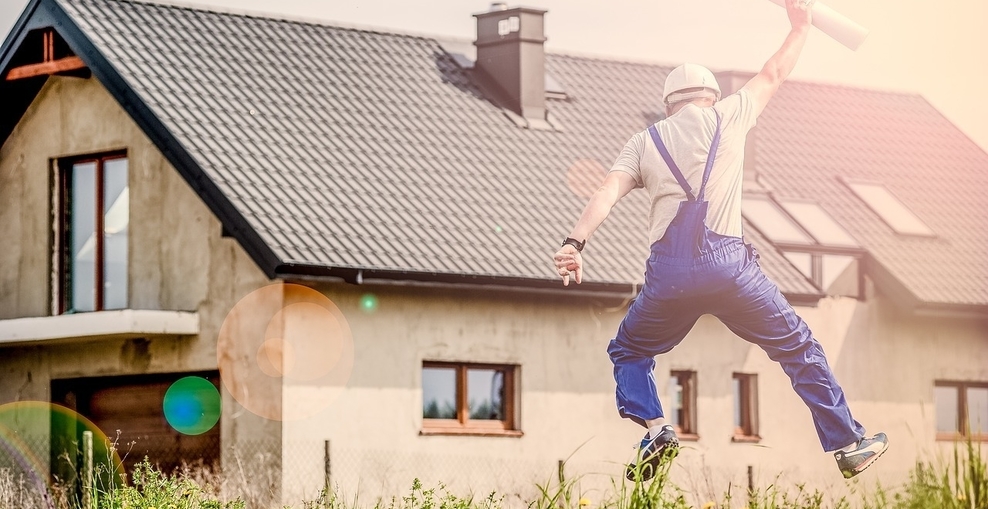 Bauarbeiter mit eingerolltem Bauplan in der Hand springt hoch, Ansicht von hinten, im Hintergrund neu gebautes Haus